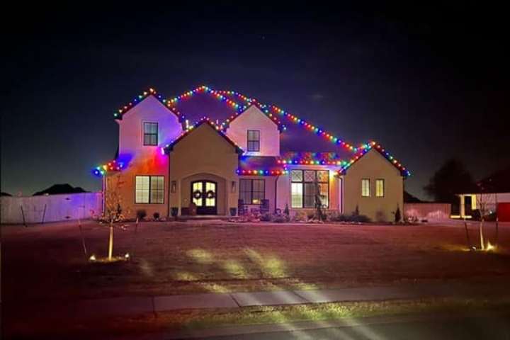 A two-story house is decorated with colorful Christmas lights outlining the roofline. A wreath hangs on the front door, and small trees with lights illuminate the lawn.