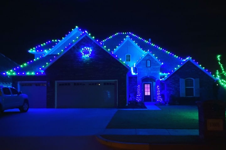 House decorated with blue and green Christmas lights. A wreath hangs over the garage, and the lawn is illuminated with green light. Holiday cheer radiates from this festive home.