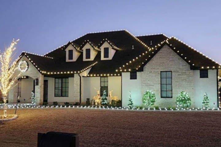 Festive home exterior at dusk, illuminated with warm white Christmas lights outlining the roof, bushes, and trees. Reindeer decorations stand on the lawn, adding to the holiday cheer.