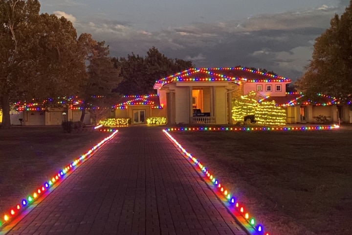 House adorned with colorful Christmas lights, outlining the roof and driveway. A bright, festive display illuminates the home and grounds, creating a warm holiday glow.