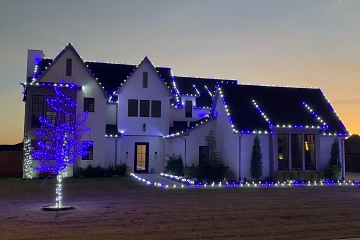 A large, luxurious home is decorated with white and blue Christmas lights outlining the roof and a blue light tree in the front yard, set against a twilight sky.