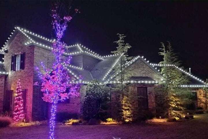 House adorned with bright white Christmas lights outlining the roofline, complemented by vibrant pink and purple lights on trees in the front yard. The festive display illuminates the night sky.