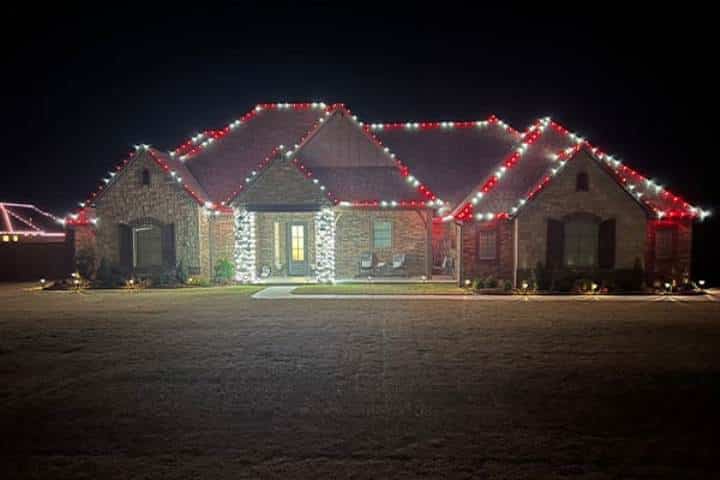 Brick house at night adorned with red and white Christmas lights along the roofline and columns. The lawn is expansive and well-lit, enhancing the festive display.