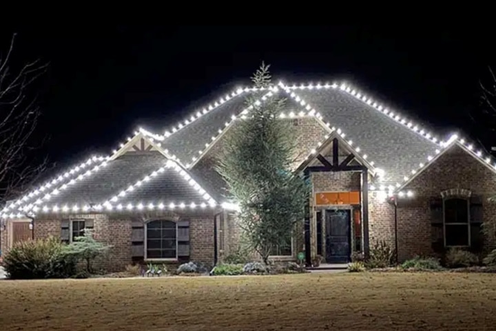 House outlined with bright white Christmas lights at night. The lights highlight the roofline and architectural details of the brick home, creating a festive and elegant display.