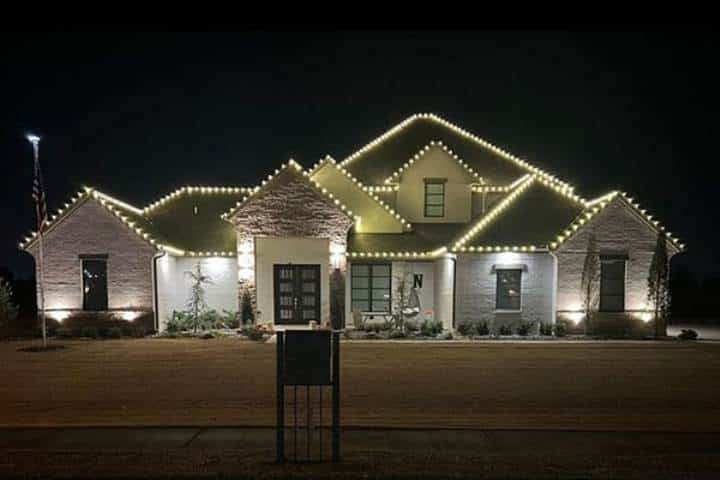 Elegant home exterior illuminated with warm white Christmas lights outlining the roofline, creating a festive holiday scene. A flag waves gently in the yard.
