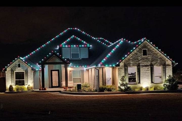 A sprawling one-story home is adorned with festive Christmas lights outlining the roofline. Red, green, and white lights twinkle against the dark night sky, illuminating the stone facade and manicured lawn.