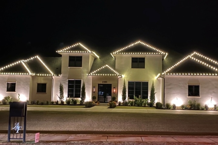 Night view of a large, stately home illuminated with warm white Christmas lights outlining the roofline. The address "2385" is visible above the dark front door.