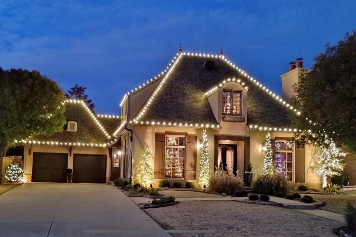 Elegant home decorated for the holidays with warm white lights outlining the roof and windows. Small, lit Christmas trees flank the front door, adding to the festive curb appeal.