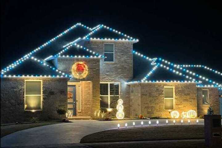 House decorated with bright white Christmas lights outlining the roof, a wreath on the front door, and glowing spherical decorations in the yard.