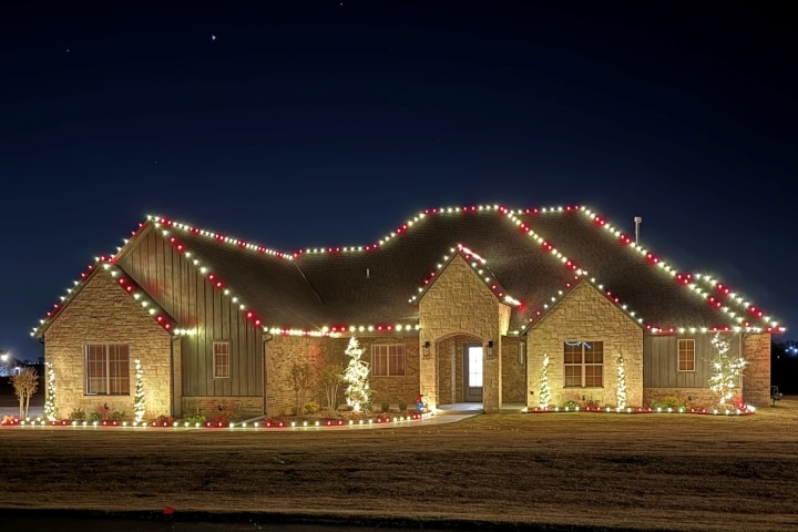 Festive house decorated for Christmas with red and white lights outlining the roof and landscaping. Warm light shines from the windows, casting a cozy glow on the lawn.