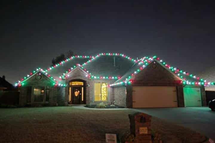 A suburban home is decorated with red and green Christmas lights along the roofline. The lights illuminate the brick facade and front yard, creating a festive holiday scene.