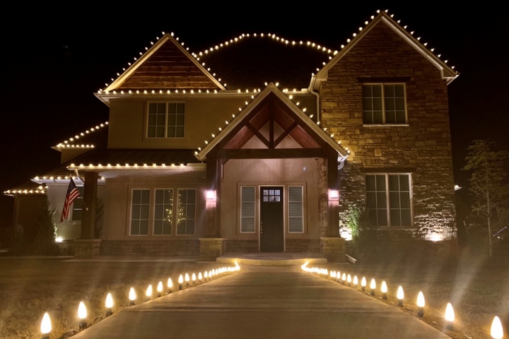 A beautiful two-story house is adorned with warm white Christmas lights outlining the roof and illuminating the driveway, creating a festive and inviting scene.